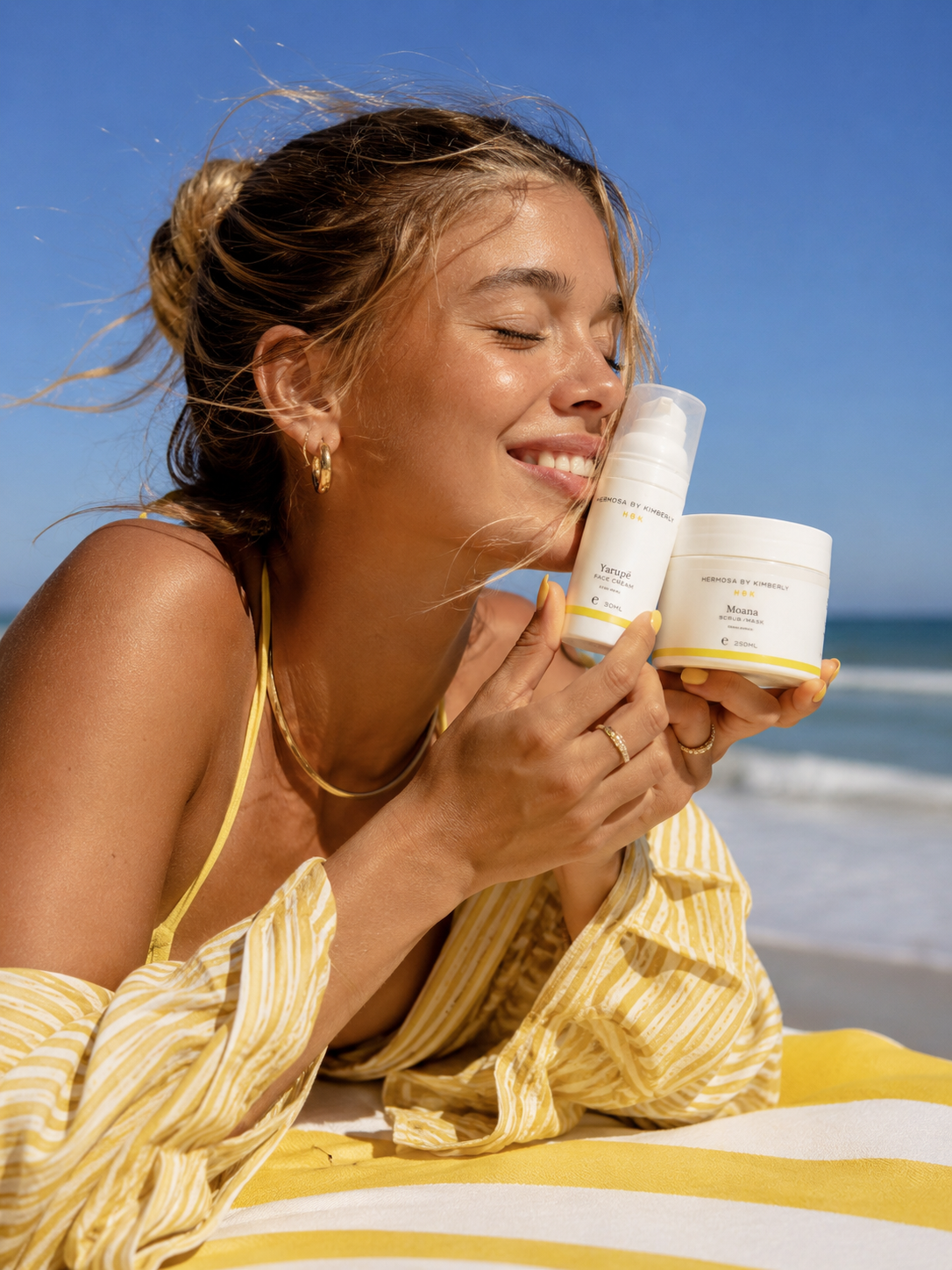 A woman lying on a beach towel at the beach, holding two skincare products from Hermosa by kimberly: psoriasis skincare/ bodycare psoriasis. huidverzorging voor psoriasis. a white pump bottle and a white jar with yellow accents.